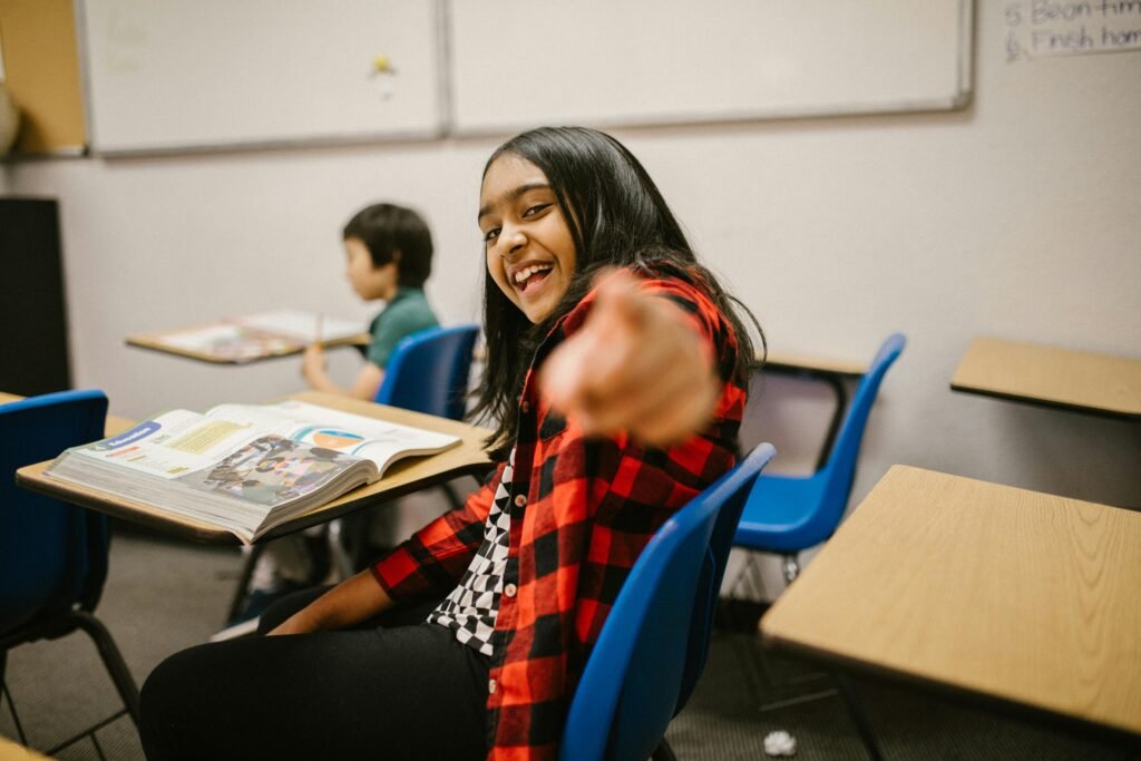 Young student in a plaid shirt smiling and pointing playfully in a classroom setting.